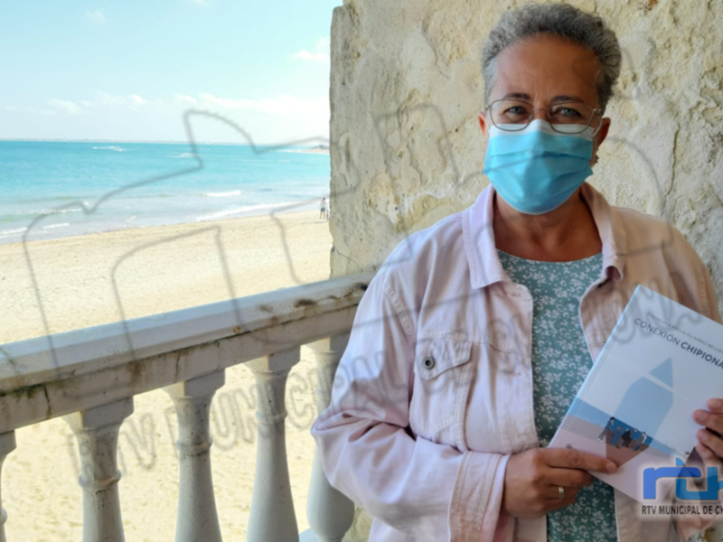 Una mujer con mascarilla sostiene un folleto en una balaustrada frente al mar.
