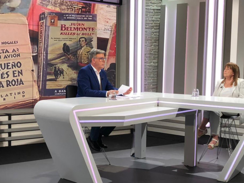 Se observa a una persona sentada en un escritorio moderno con líneas de luz LED, frente a un fondo de libros y una pared de ladrillos. La persona parece estar en un estudio de televisión o presentando algo, lo que sugiere una escena relacionada con la cultura o el entretenimiento.