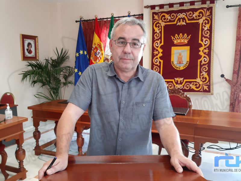 Un hombre en una oficina, frente a una bandera española y una bandera europea, sosteniendo una pluma y escribiendo en un papel. El edificio es de estilo clásico con muebles de madera y una bandera municipal en el fondo.