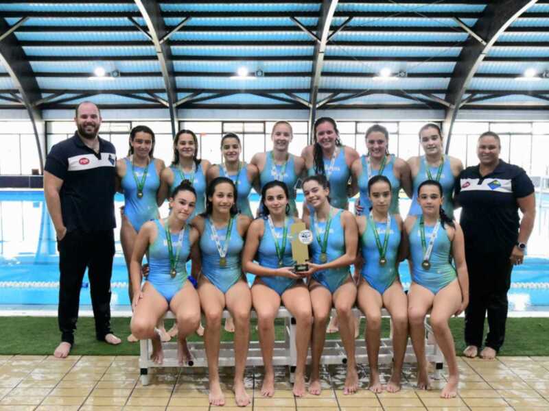 Equipo femenino de natación posando con sus medallas y trofeo en la piscina.