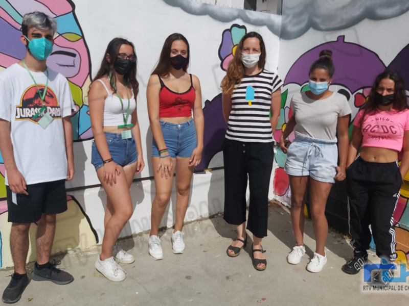 Estudiantes posan frente a un mural con personajes de dibujos animados, todos con máscaras faciales. La imagen muestra una escena casual y colorida en un entorno urbano.