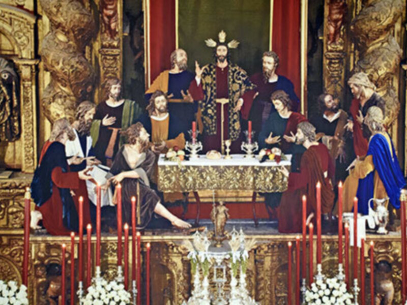 Escena religiosa con personajes vestidos en trajes históricos, posiblemente de la Edad Media. La figura central parece ser un monarca o líder religioso, rodeado por otros personajes. La decoración incluye velas y adornos dorados en un entorno de paredes ornamentadas.