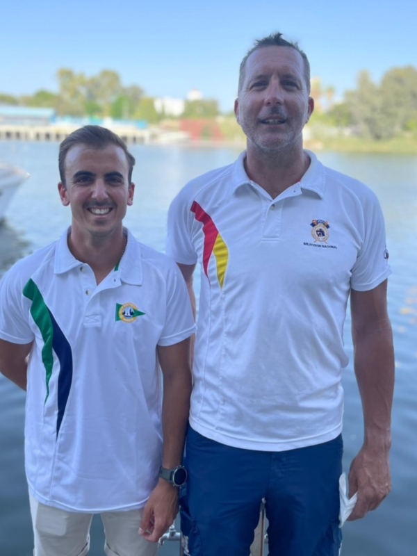 Dos hombres en camisas blancas con insignias nacionales posan en un muelle con agua azul y cielo claro al fondo.
