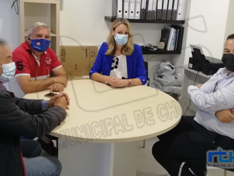 Una reunión en una oficina con cuatro personas sentadas alrededor de una mesa. La imagen muestra a dos hombres y una mujer en la mesa, con un hombre de pie detrás. Todos están usando máscaras faciales y hay documentos y carpetas en la pared de fondo. La imagen está firmada con "MUNICIPAL DE CHIPONA" en la esquina inferior derecha.