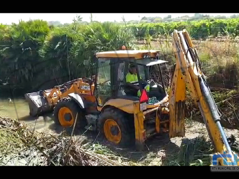 Un excavador amarillo trabaja en un canal, con vegetación y agua alrededor. El operador viste una chaqueta verde.