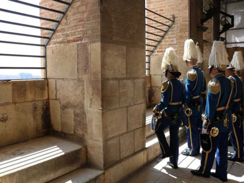 Guerreros en uniforme azul y dorado, posando en una escalera de mármol, con un fondo de una pared de ladrillos y un ventanal con persianas negras. La escena transmite una sensación de solemnidad y formalidad.