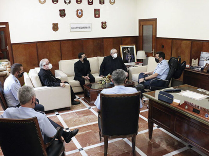 En una sala de conferencias, un grupo de personas se reúne en torno a una mesa redonda. La pared está decorada con medallas y insignias, y hay un escritorio en la esquina. La atmósfera parece formal y seria.