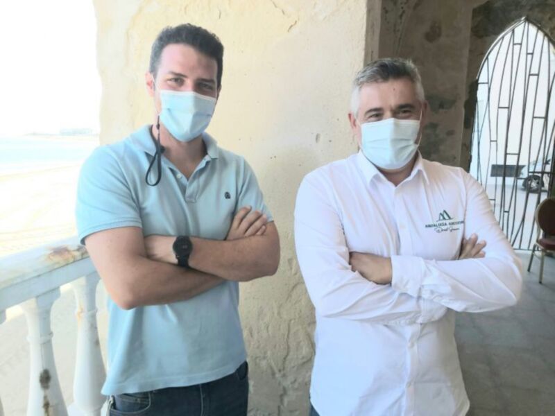 Dos hombres vestidos con ropa casual y máscaras faciales, posando en un entorno de exterior con una estructura de piedra y una puerta metálica en el fondo.