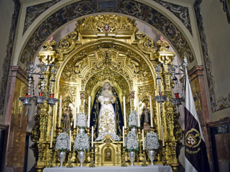 Interior de una iglesia con una imagen de la Virgen María en el altar principal, rodeada de adornos dorados y una bandera en la pared.