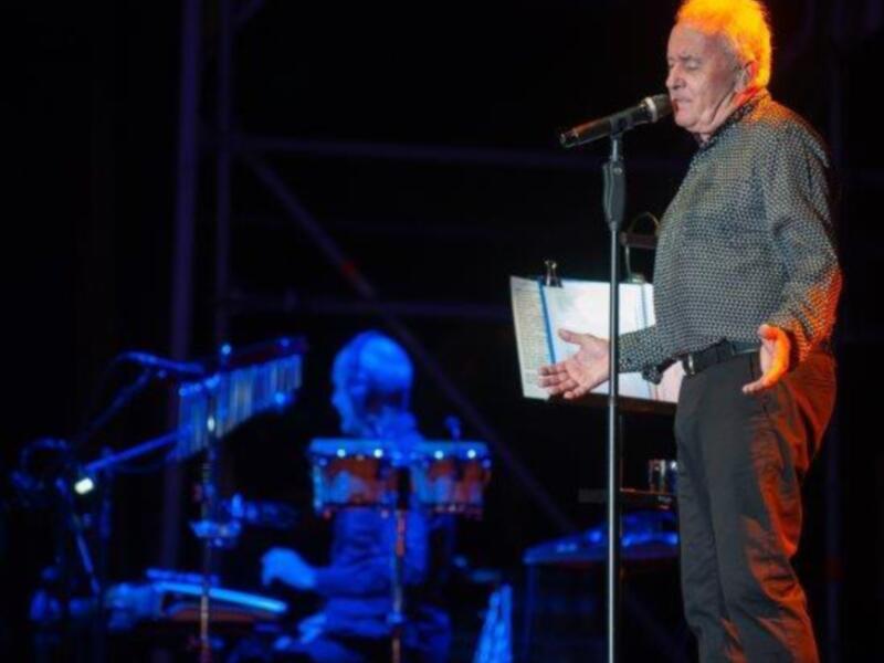 Un músico en un escenario de concierto, con un piano y un segundo músico en el fondo, iluminado por luces azules y rojas.