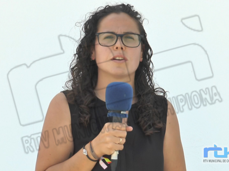 Una mujer con cabello rizado y gafas sostiene un micrófono azul, posiblemente en una entrevista o reportaje. El fondo es claro con un logo y texto que parece ser de una emisora local.