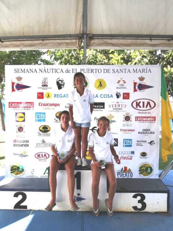 Tres jóvenes ganadoras posan en el podio de la Semana Náutica de El Puerto de Santa María, con sus trofeos y sonrientes. El fondo muestra logos de varias empresas patrocinadoras de la competición.