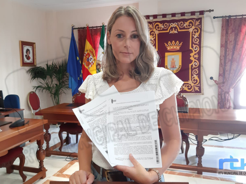 Mujer en oficina municipal con documentos, fondo con banderas y escudo de Chipiona.