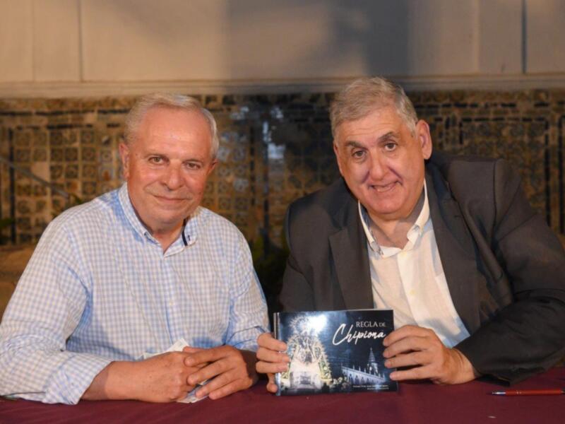 Dos hombres sonrientes, uno en camisa a cuadros y el otro en traje, sostienen un libro titulado 'Regla de Chipiona'.