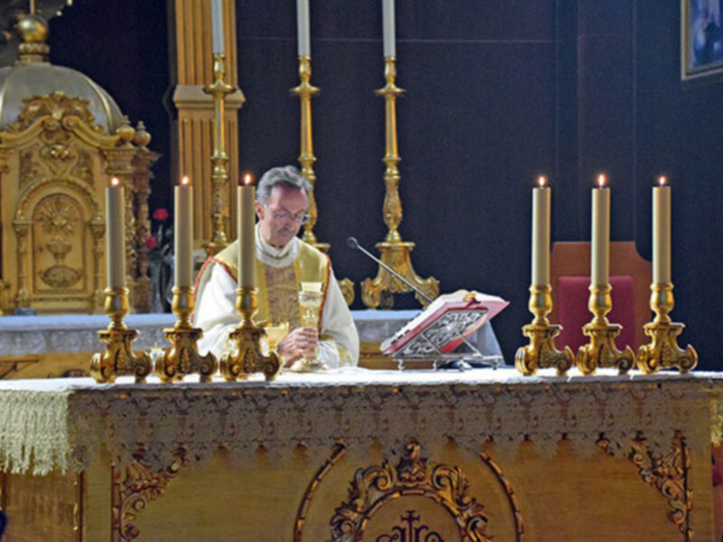 Un sacerdote vestido con un hábito blanco y dorado, sosteniendo una copa de vino mientras lee un libro en el altar. El altar está decorado con velas doradas y una base de madera tallada, en un entorno religioso.