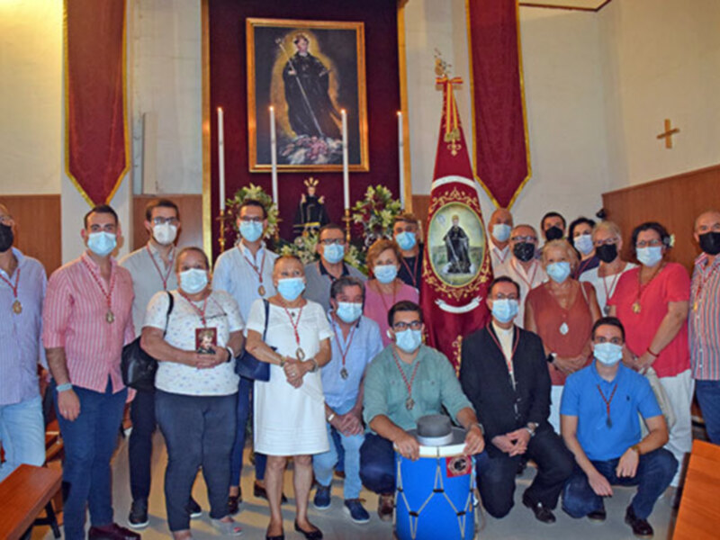 Grup de persones amb mascaretes en un ambient religiós, probablement dins d'una església. Al fons, hi ha una imatge de la Verge Maria amb un escut i creus. Les persones porten roba formal o casual, alguns amb llancetes. La imatge transmet una celebració o assemblea religiosa.