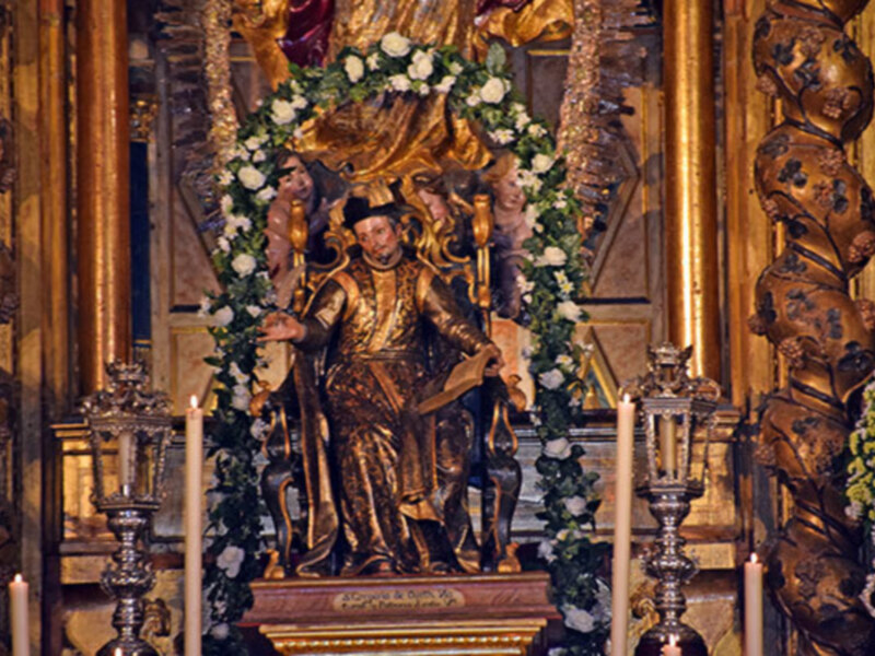 Estatua de un personaje histórico vestido con trajes dorados, sentado en un trono, rodeado de flores y velas en un entorno de madera tallada.
