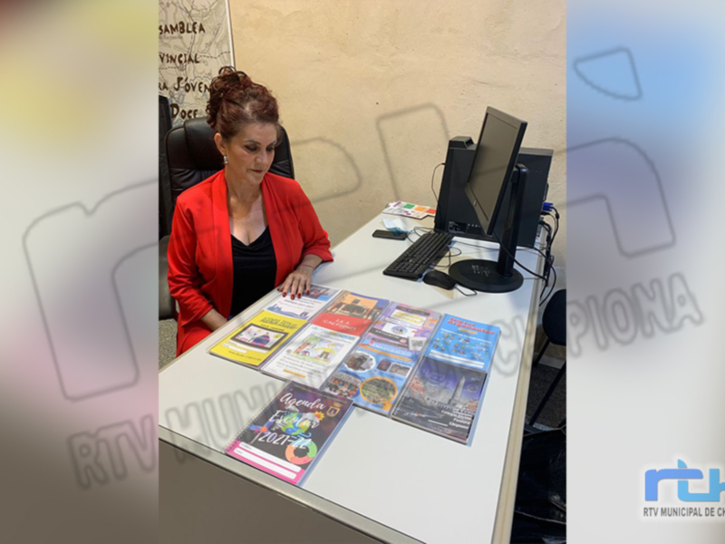 Una mujer sentada en una mesa frente a un ordenador, con varios libros de color y texturas diferentes sobre la mesa. La imagen parece estar en un entorno profesional, posiblemente una oficina o biblioteca.