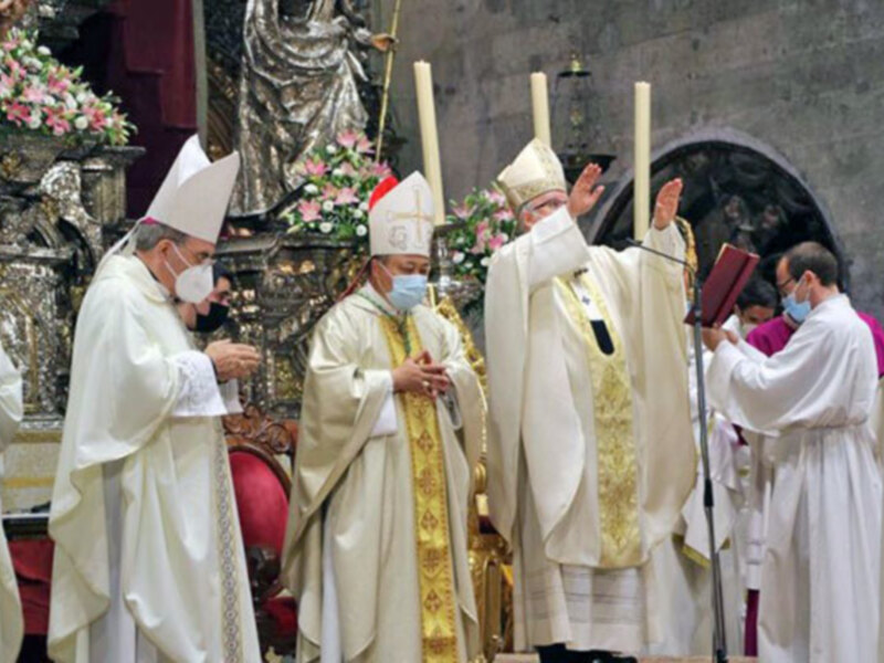 Imagen de una procesión religiosa con sacerdotes vestidos con hábitos blancos y dorados, usando máscaras faciales, frente a una estatua de una figura religiosa.