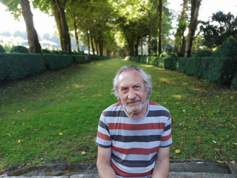 Un hombre con barba blanca y una camiseta a rayas sentado en un parque con árboles y césped.