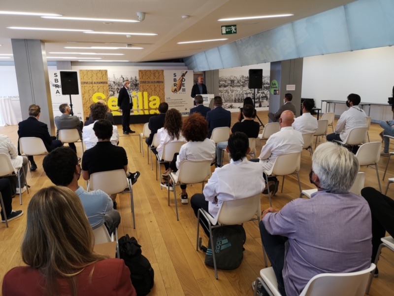Sala de conferencias con asistentes sentados en sillas alineadas, frente a una mesa de conferencia con un panel de presentación. La sala tiene iluminación artificial y paneles de madera en el fondo con texto visible.