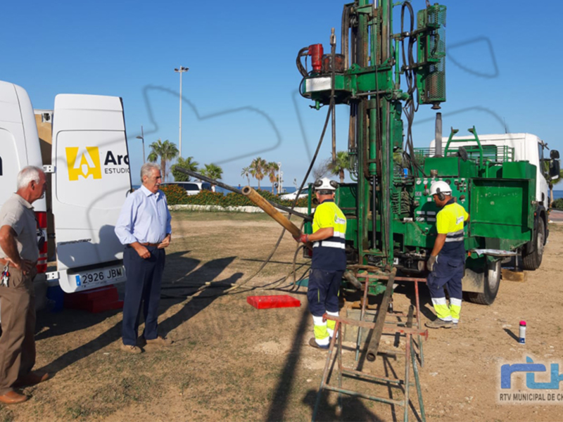 Trabajadores de una empresa de servicios públicos operan una máquina de perforación en un área de construcción. En el fondo, se pueden ver camiones y una carretera con árboles a lo lejos.