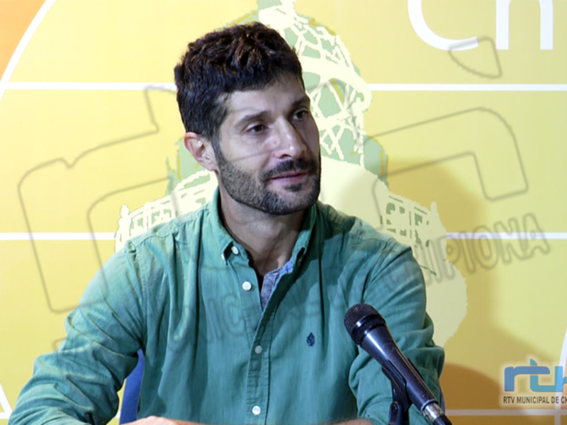 Un hombre con barba y cabello oscuro, vestido con una camisa verde, está sentado en frente de un micrófono. El fondo es amarillo con una imagen de un león y el texto "RTV.MUNICIPAL.DE.CHIPIONA" en la esquina inferior derecha.