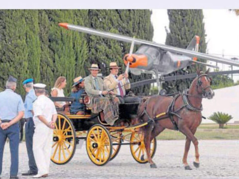 Un avión de ala fija sobrevuela una carroza tirada por un caballo, mientras una multitud de personas observa la escena.