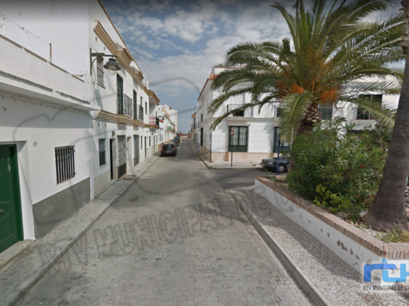 Calle estrecha con casas blancas y un árbol de palma en el lado derecho, en un entorno urbano con cielo azul y nubes dispersas.