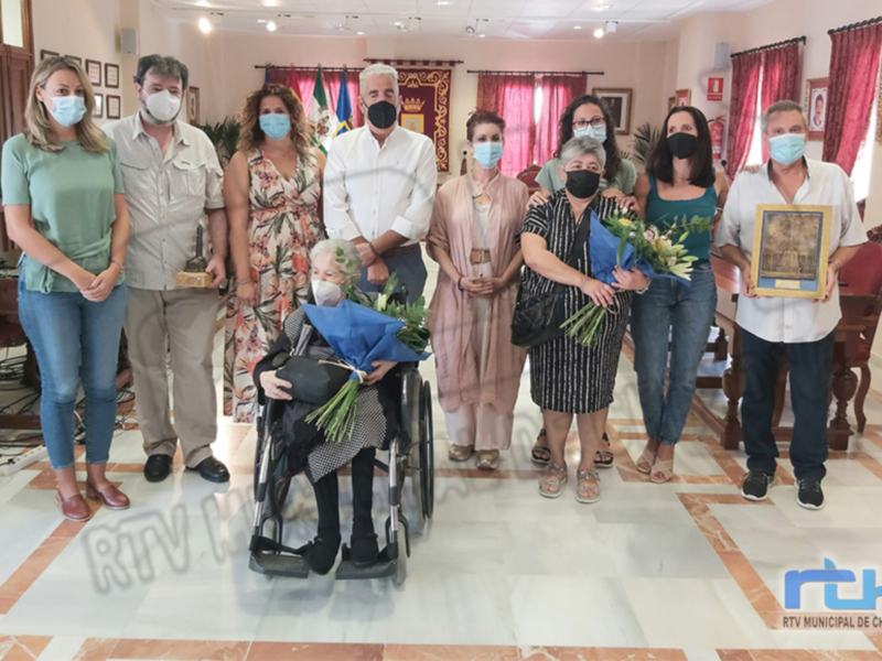 Una imagen de una reunión formal en un salón, con siete personas posando para la foto. La persona central está en una silla de ruedas, y todos están usando máscaras. La imagen muestra un ambiente formal con decoración y una mesa de centro visible en el fondo.