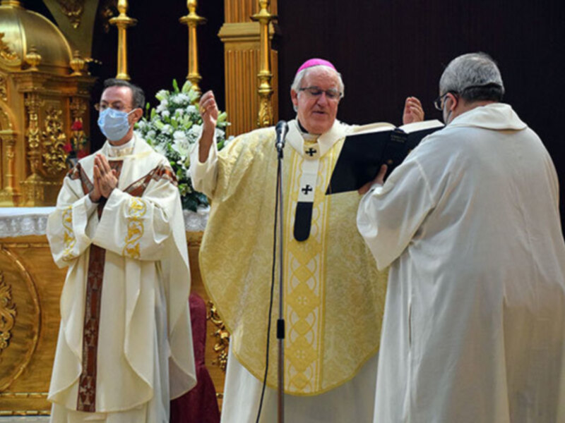 En una ceremonia religiosa, tres sacerdotes vestidos con vestiduras blancas y doradas se encuentran en un altar elaborado. El sacerdote central, con una cruz dorada en su chasuble, realiza gestos de oración mientras el sacerdote izquierdo, con una máscara médica, y el derecho observan atentamente. La imagen transmite un ambiente serio y reverente típico de una celebración religiosa.