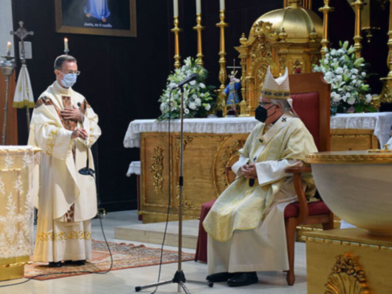 Imagen de dos sacerdotes en una iglesia, uno con mascarilla y el otro sentado. El altar está decorado con flores y la imagen del Cristo en el fondo.