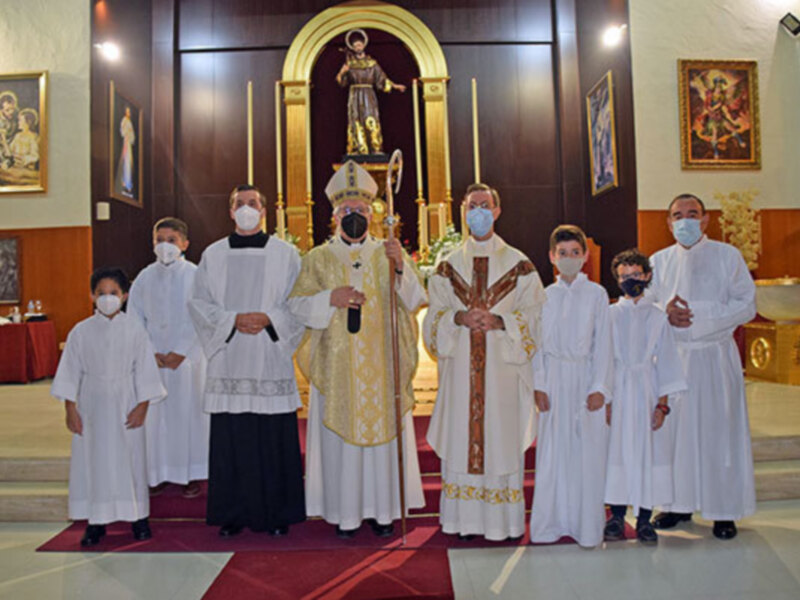 Imagen de una iglesia donde se observa a varios sacerdotes vestidos con hábitos blancos y otros vestidos de manera formal. En el fondo, se puede ver una estatua religiosa y varios cuadros decorativos. La imagen parece ser tomada durante una ceremonia o celebración religiosa, con todos los presentes usando mascarillas.