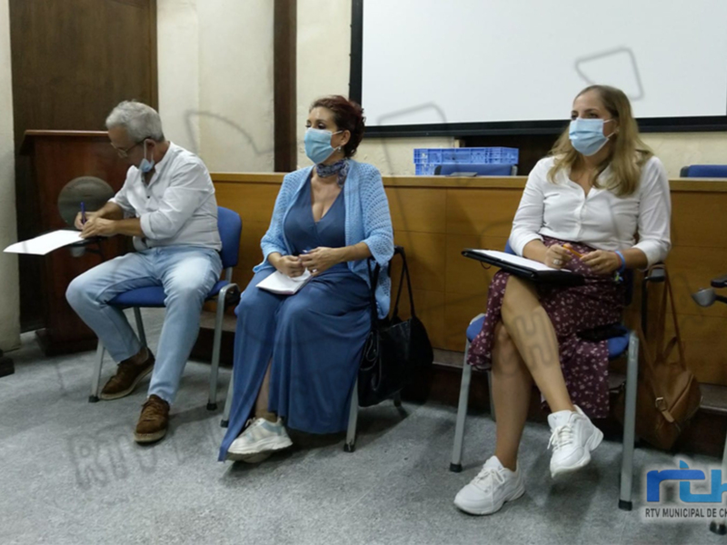 Tres personas sentadas en una sala de reuniones, con máscaras faciales, participando en una discusión o reunión formal.