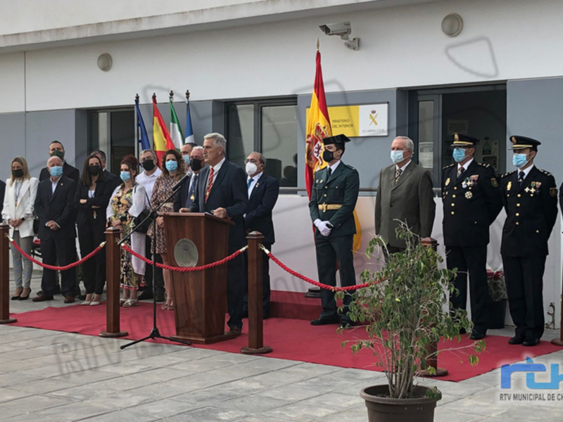 Evento oficial con personas en trajes formales y máscaras, frente a una bandera y un edificio blanco.