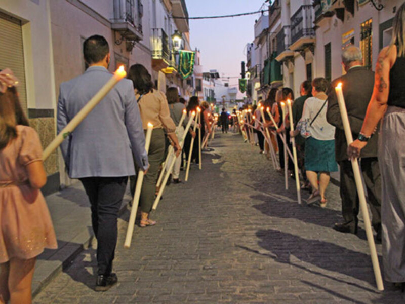 Una procesión religiosa con personas sosteniendo bastones de madera iluminados por velas en una calle estrecha y adoquinada.