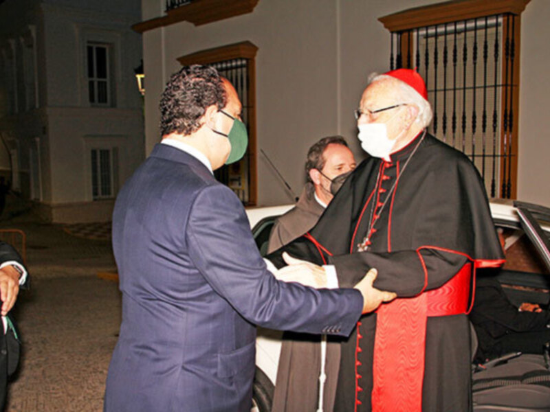 Un hombre en traje azul se despidiendo de un hombre en traje negro con capa roja, ambos con máscaras médicas en un entorno urbano nocturno. La escena parece ser en un evento formal o ceremonial, con un vehículo blanco y una puerta de madera en el fondo.
