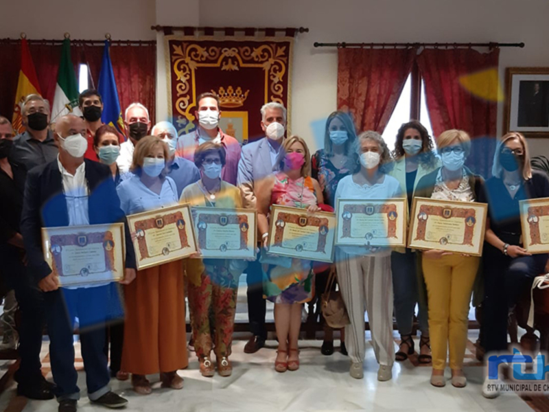 Una foto de grupo en un entorno formal, con varias personas vestidas formalmente y sosteniendo diplomas. La imagen muestra a una persona en el centro con un vestido colorido, y alrededor hay personas con diferentes estilos de ropa. La escena parece ser en una sala de conferencias o oficina, con banderas y decoración en el fondo.