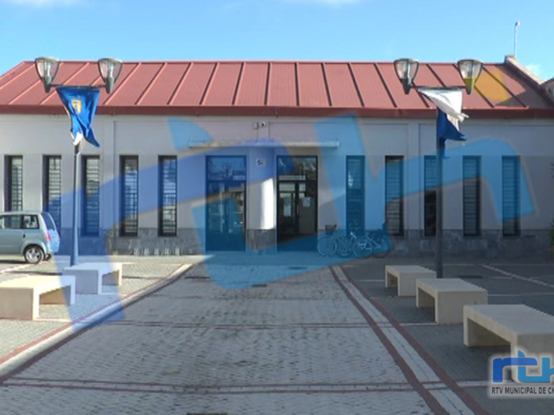 Una vista de un edificio gubernamental con una cubierta roja y ventanas azules. Hay dos bicicletas aparcadas frente al edificio, un coche gris estacionado a la izquierda y una bandera ondeando en el lado derecho. El cielo es azul con algunas nubes dispersas.