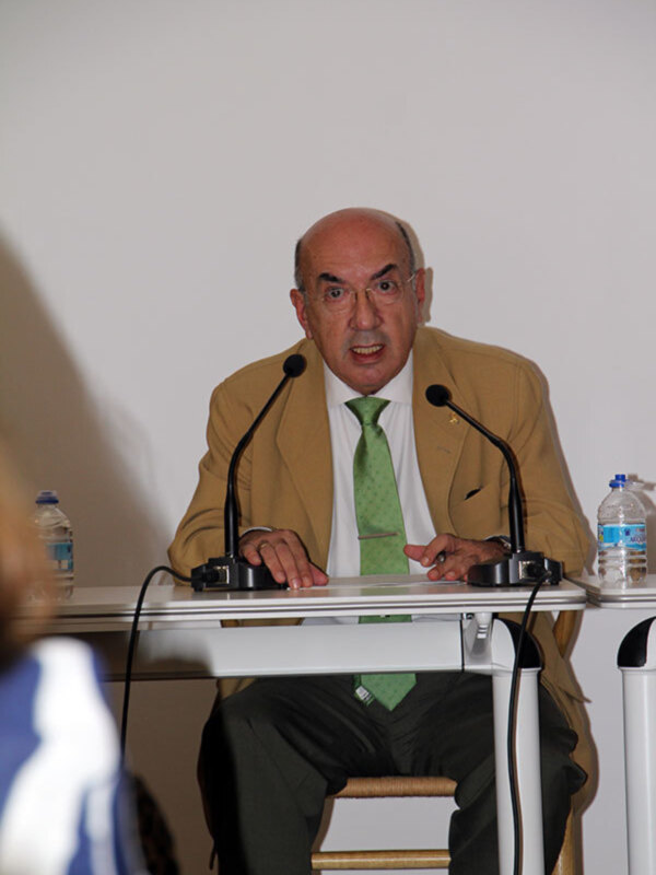 Un hombre en un traje beige y corbata verde está sentado en una silla de madera, hablando en un micrófono. En la mesa delante de él hay una botella de agua y un portapapeles. La pared al fondo es blanca.