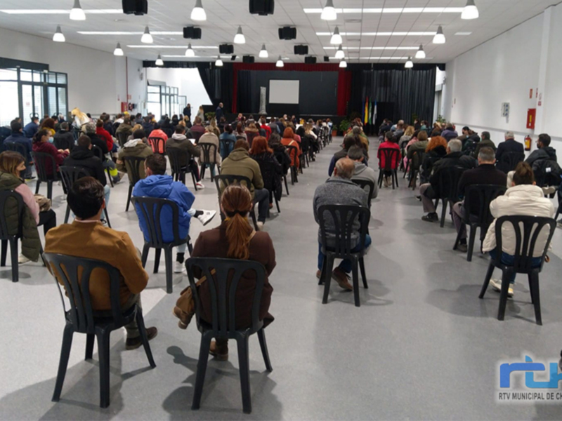 Una sala de conferencias con una audiencia sentada en sillas, frente a un escenario. La sala está iluminada con luces de techo y hay cortinas negras en el fondo. La imagen muestra una conferencia o presentación formal.