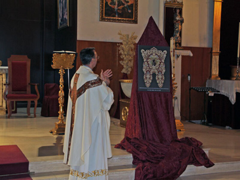 Un sacerdote en vestimenta litúrgica se encuentra en una iglesia, con un altar y una silla roja al fondo. La imagen muestra detalles de la decoración religiosa, como un tapiz y una lámpara dorada.