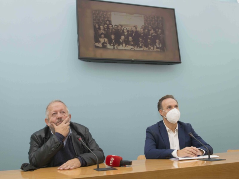 Dos hombres sentados en una mesa de conferencia, uno con una máscara y el otro sin ella. En la pared detrás de ellos se puede ver un cuadro con una foto de grupo.