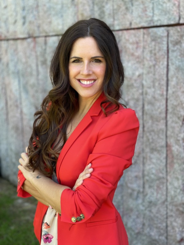 Elegante mujer con blazer rojo y blusa floral, posando frente a una pared de piedra.
