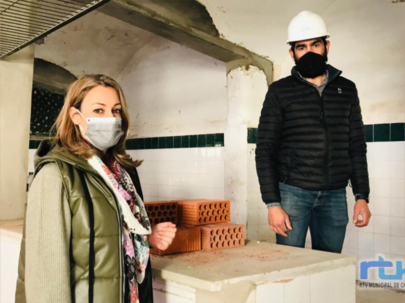 Dos personas con cascos de construcción y mascarillas, posando frente a un muro de ladrillos.