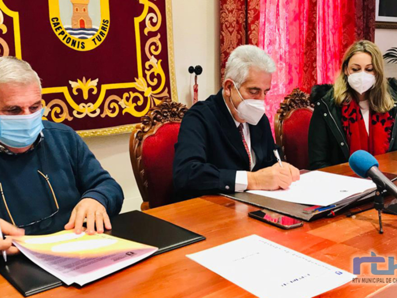 A tres personas sentadas en una mesa de conferencia, firmando documentos. La persona central lleva un pañuelo facial y está sosteniendo un documento mientras firma. A su izquierda, otra persona también lleva un pañuelo facial y sostiene un documento. A la derecha, una tercera persona con cabello largo lleva un pañuelo facial y sostiene otro documento. La mesa tiene varios documentos y una pluma en ella, y el fondo muestra un escudo con texto que parece ser la bandera de una institución.