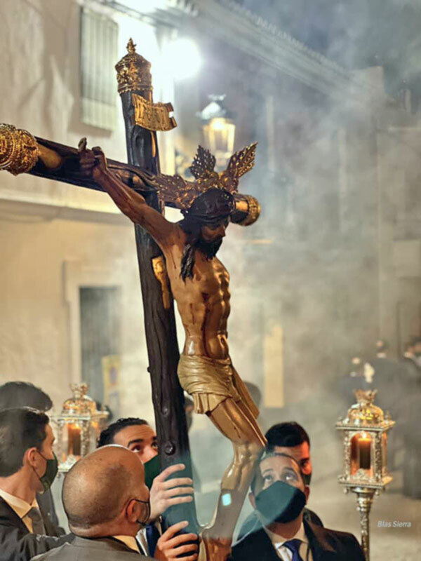 Estatua de Cristo crucificado en una procesión religiosa, con personas vestidas formalmente y luces de velas en el fondo.