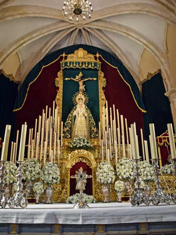 Un altar de iglesia con una Virgen María en el centro, rodeada por flores y velas. El techo tiene una arquería gótica y una lámpara de cristal colgante. La decoración es dorada y azul, con detalles en oro y azul marino.
