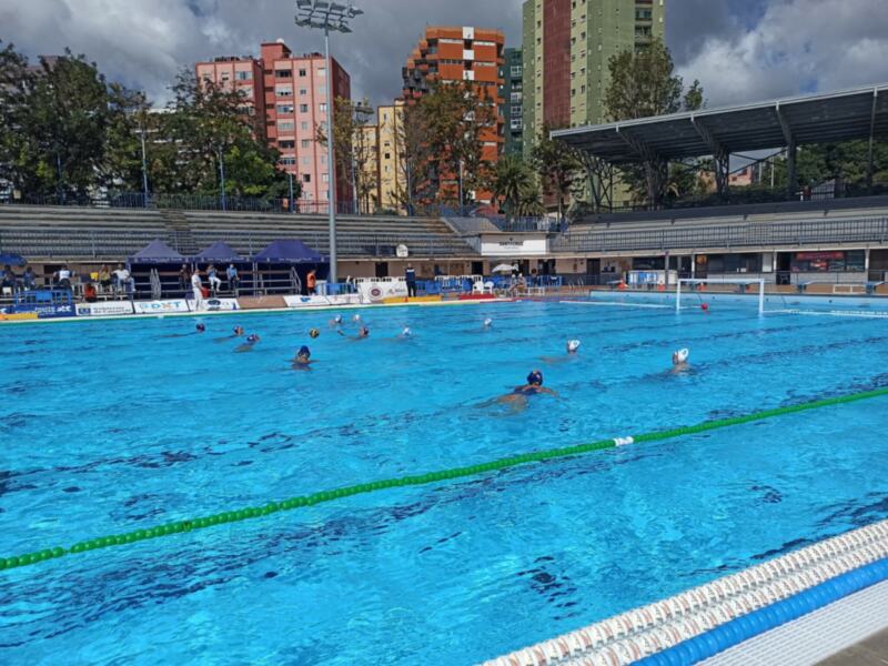 Jugadores de waterpolo compiten activamente en una piscina al aire libre, rodeada por edificios urbanos.