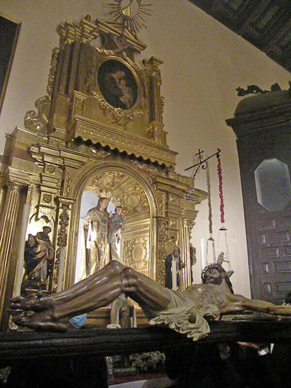 Un altar de estilo barroco con una escultura del Cristo Crucificado en el centro. Alrededor, hay estatuas de santos y una imagen de la Virgen María en un marco dorado. La pared está decorada con detalles dorados y una puerta de madera con un espejo octogonal.